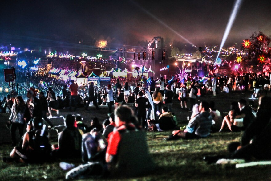 Crowds of people are sitting on a grassy slope at night during a large music festival. Lights, stages, lasers, and vendors can be seen in the distance. Even more people are walking around the event grounds in the background.