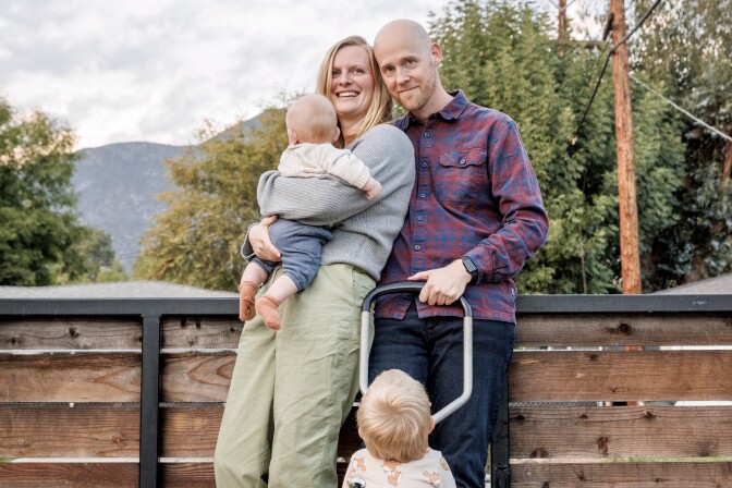 A white couple stands outdoors in front of a wooden fence, smiling at the camera. The woman holds a baby. A toddler stands in front of them with their back to the camera.