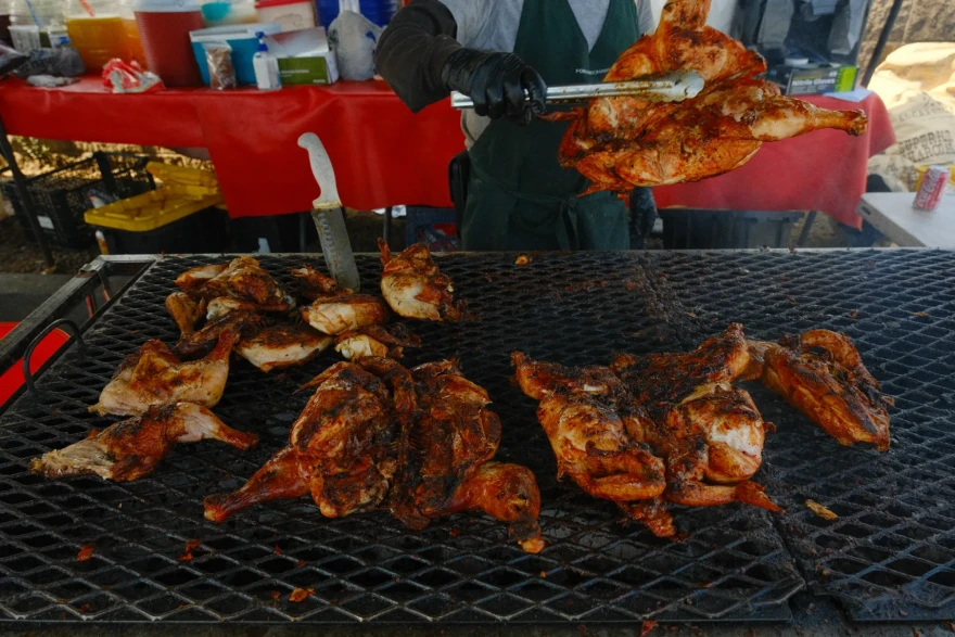 A vendor grills and flips seasoned chicken over an open flame at an outdoor food stand, with various supplies and drinks in the background.