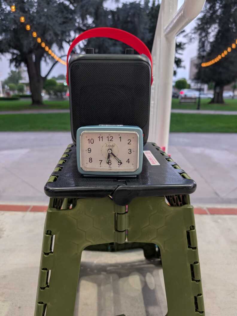 A clock and an audio speaker / CD player on top of a stool at a park