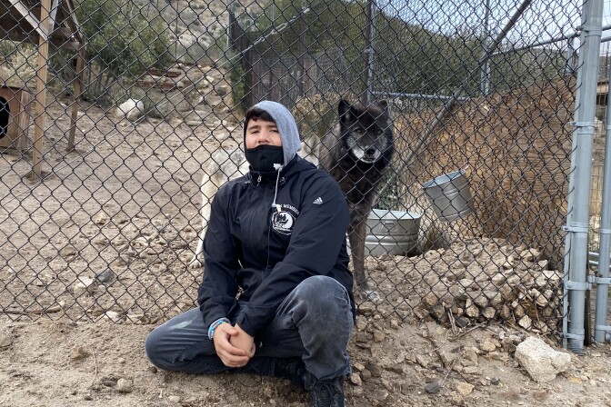 Wolf Connection team member Edward Amaya sits with hands clasped together. He wears a black jacket and grey hoodie. Beside him, behind a fence, sits his buddy Kenai, a black and brown male wolf who lives on the ranch. 