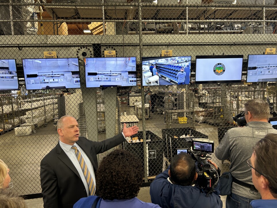 A man in a suit gestures toward a line of monitors attached to a chain fence showing different aspects of ballot counting and other election operations. 