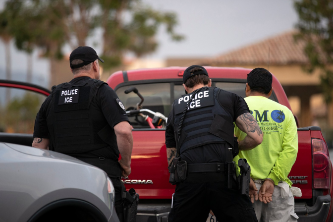 Two police/ICE officers in front of a red truck arresting something in a bright yellow shirt