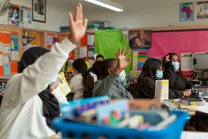 A group of children sitting at their desks raising their hands. There is a blurry blue bucket with school supplies at the front center of the photo. In the background are colorful papers hanging from the wall.