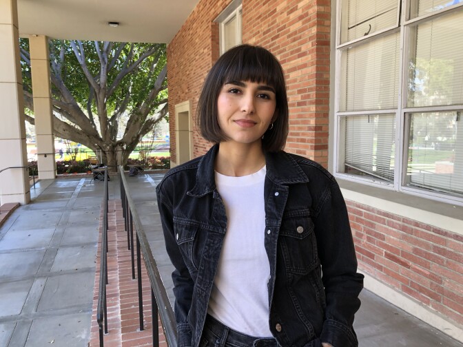Woman in her 20s with bob haircut and black denim jacket stands next to brick wall
