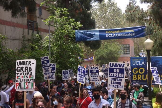 About two dozen academic workers are gathered outside a university campus with signs that say "UAW. On Strike. Unfair Labor Practice."