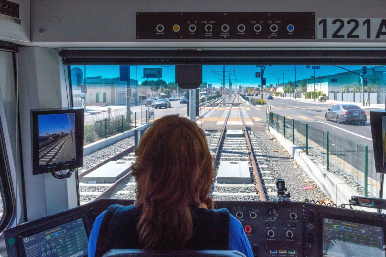 A person sits at a control panel with their back to the camera as we see train tracks running in the middle of a main roadway out the front window.