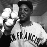A man is holding up four baseballs in one hand. He's smiling and wearing a San Francisco jersey. 