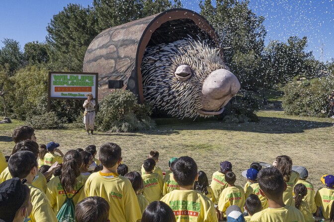 A massive porcupine puppet head peaks out of a faux log as kids watch nearby.