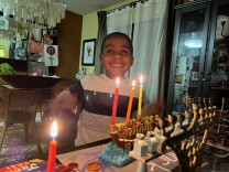 A young boy in a white and black block-print sweater sits at a table behind the lit candles of a menorah.
