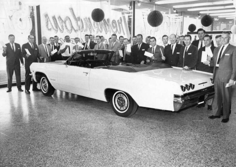 men holding up checks stand around a white car inside an auto dealership.