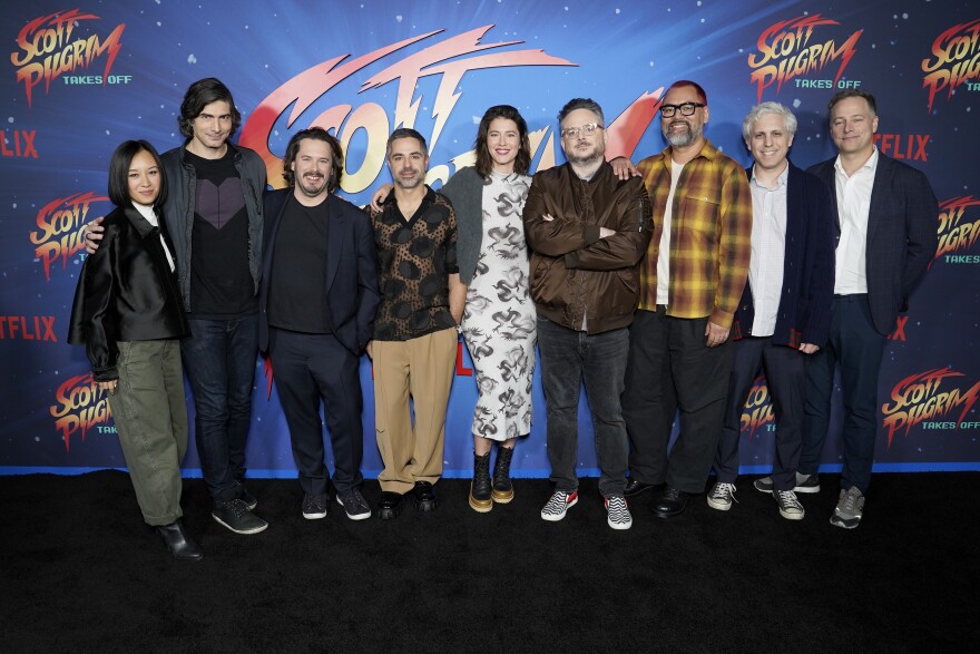 Nine people stand side-by-side, behind them ifs a backdrop for the Netflix program "Scott Pilgrim Takes Off"