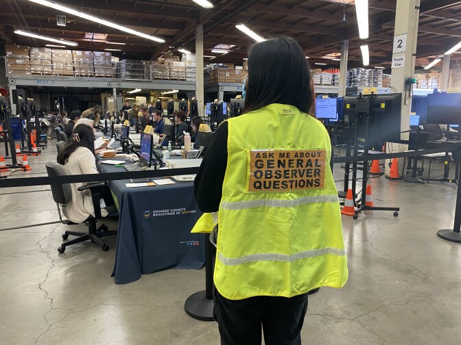 In the foreground, a woman with her back turned wears a bright yellow vest with a sign that reads, "Ask me about general observer questions." In the background, a row of people sit in front of computers. 