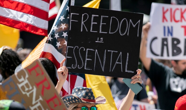 A person wearing a hat with U.S. flag-style stars holds up a sign that reads: Freedom is Essential