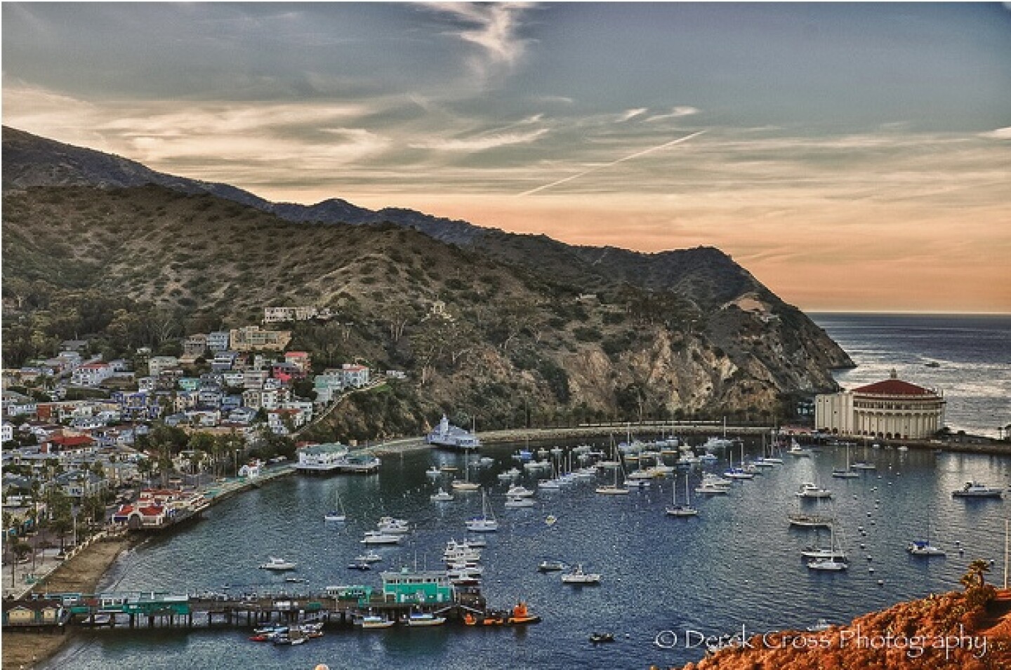 An orange-blue sunset is pictured over the hills and bay of Catalina Island. Many sailboats dot the water and houses are seen on the hill to the left.