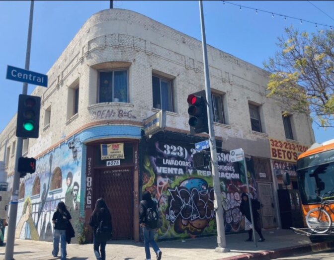 A white historic corner two story building sits with traffic lights either side and a sign that says Central Avenue. It has advertising posters on its first floor