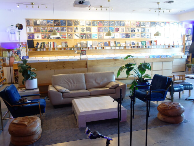Lounge area in a record shop with a gray sofa, two blue armchairs, and a low pink table on a patterned rug, with album covers displayed on the wall behind and potted plants nearby.