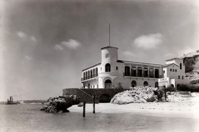 A black and white photo depicts a beachfront marine lab with a central tower and tiled roof. 
