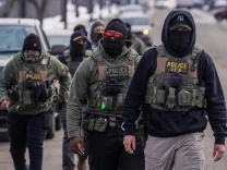 Federal agents, wearing face masks, vests, and some wearing eyewear, walk down a street.