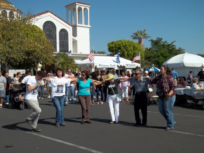 A group of perform a traditional Greek golk dance in a Church parking lot.