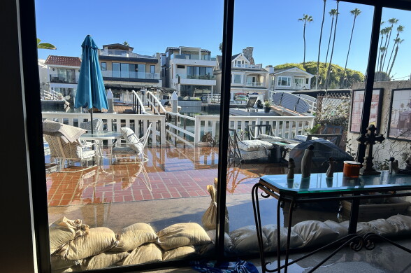 Sandbags line a glass door as floodwater covers a patio along a waterway lined with homes and docked boats.