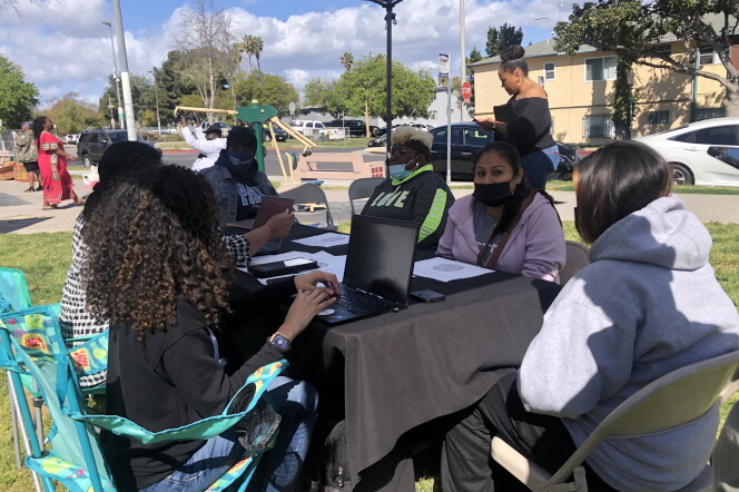 A group of women gathered around a table under a canopy. The table has a black tablecloth. Two of the women work at laptops, others wearing masks. Other women walk by in the background. They are sitting in chairs on grass, with a residential neighborhood behind.