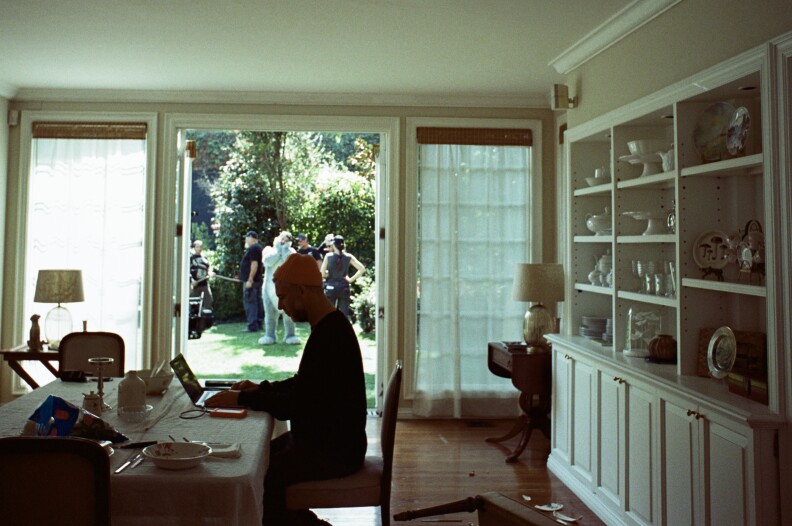 A person types on a small laptop at a desk wearing a knit cap. The room has open bookshelves with glassware. A view through French doors show a small group clustered on the lawn.