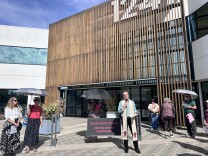 A man wearing a cream-colored stole speaks into a microphone in front of a building. A group of people stand in a semi-circle around him, several holding umbrellas to shield them from the sun. 