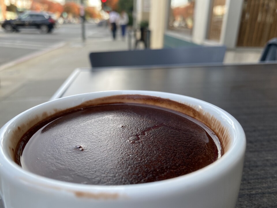 A photograph of a cup of hot chocolate sitting in a white mug placed on an outdoor patio tabletop. The chocolate is so rich the surface of the drink is leaving thick chocolate-y marks in its wake. 