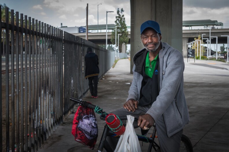 A Black man wearing a blue baseball cap, an unzipped gray sweater, and a green polo shirt, sits on his bicycle seat. A plastic bag and other belongings hang from the handlebars. To his left is an iron fence, and behind him is a cement pillar. Another man with his back turned leans against the fence in the background.