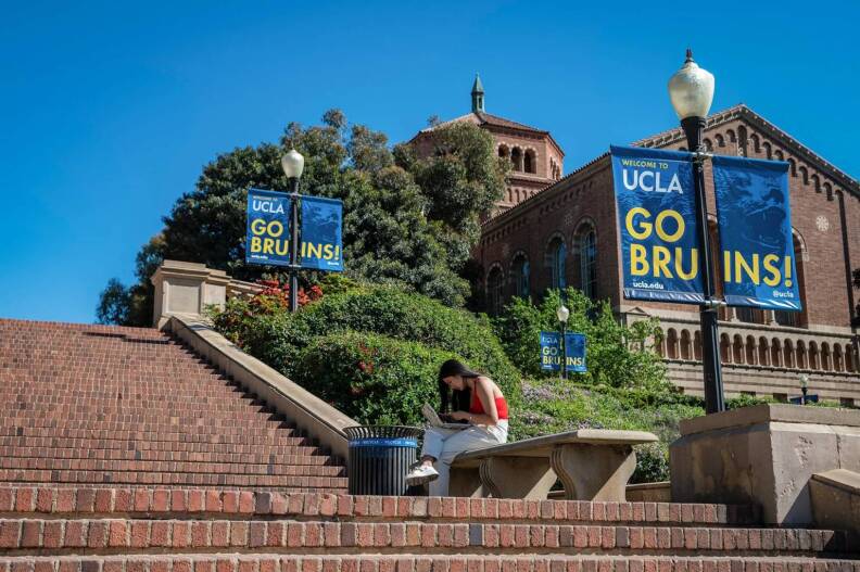 A student sits on a bench along a brick stairway outside. Light posts are set along the stairway with signs that read "Welcome to UCLA. Go Bruins!" Trees and a brick building are in the background.