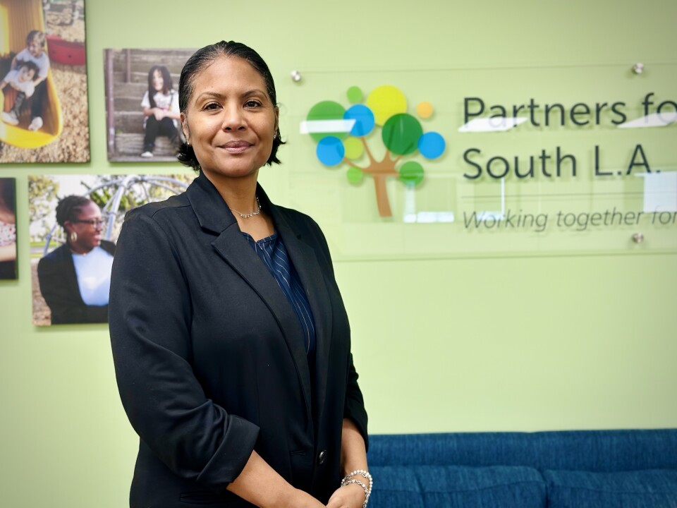 A medium skin-toned woman in a black suit stands in front of a colorful Partners for Children South L.A. sign, smiling and looking into the camera confidently.