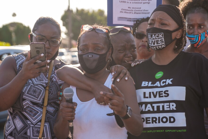 Several Black people are seen at a protest against the shooting of Dijon Kizzee by a sheriff's deputy. One woman is holding a smart phone, another woman in a white tank top is holding a microphone. A woman wears a mask that reads BLACK LIVES MATTER and a t-shirt that reads BLACK LIVES MATTER PERIOD.