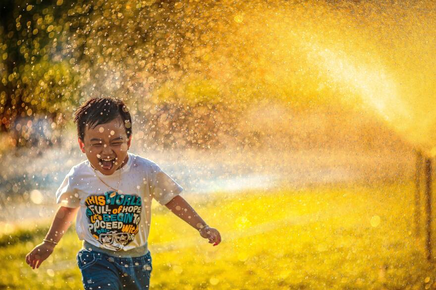 A young Asian child runs through a sprinkler wearing a t-shirt and jeans. He is laughing and the sunshine is bright behind him, illuminating the water. 