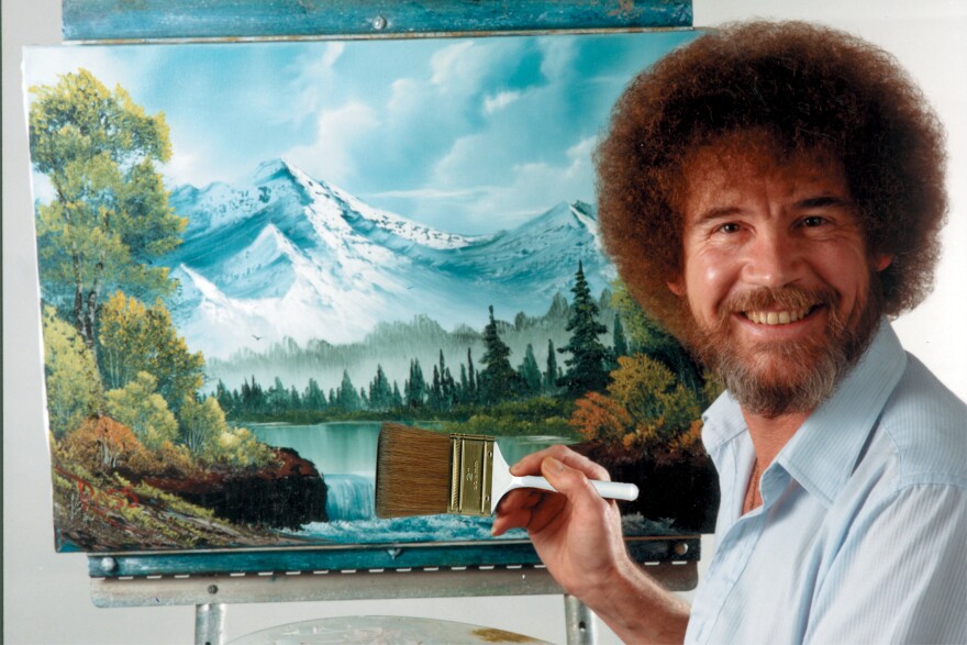 A man with a beard is smiling, wearing a blue collared shirt and holding a paintbrush in front of a landscape painting