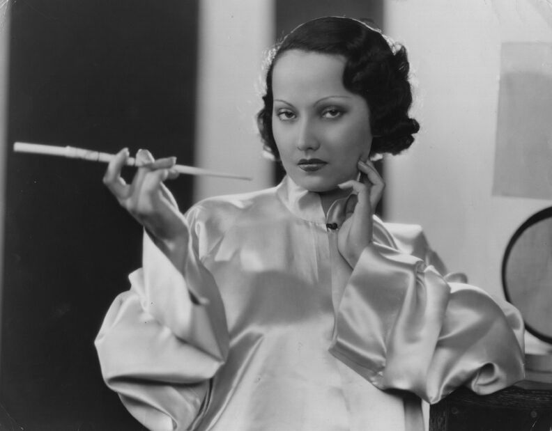 A woman in a silky blouse holds a cigarette holder. She gives a cold stare to the camera. She has black hair.