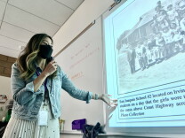 An Asian American woman with ombre dyed hair and wearing a jean jacket stands before a white board onto which a black-and-white photo of Japanese American schoolchildren has been projected. 