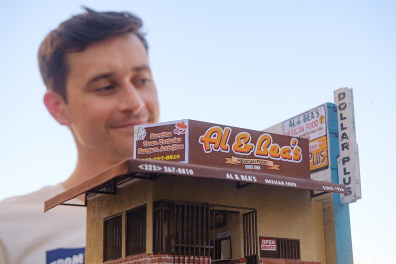 A small version of Al & Bea's Mexican Food in the foreground. It's a brown and tan-colored building with an awning that overlooks a front entrance with metal bars. A small open sign is in the window. On the right side is a wall that has a sign for the business and a Dollar Plus store. The building is being held up by the artist, who is standing in the background.