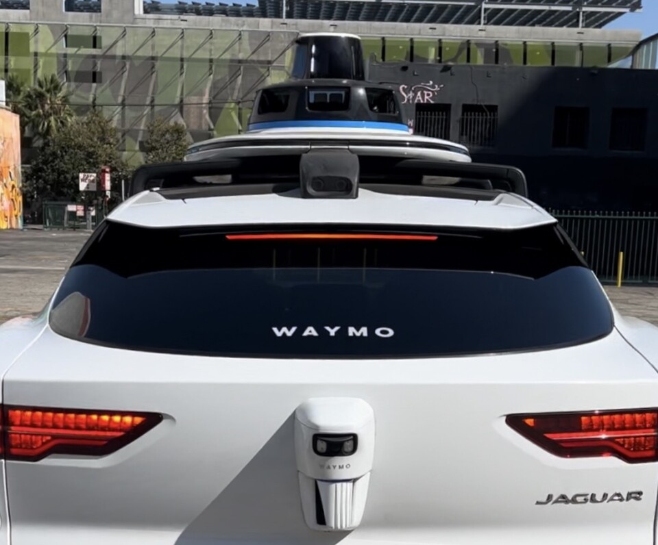 The back of a white Jaguar, with the word "Waymo" in white in the middle of the back window. 