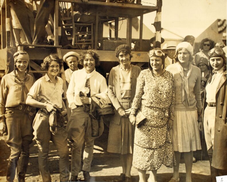 A sepia toned photograph shows a group of women in different outfits from the 1930s standing outside in a line smiling at the camera