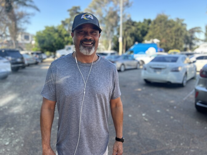 An older Black man with a grey mustache and beard and a blue baseball smiles and wears a grey workout shirt. In the background are cars parked in a parking lot. 