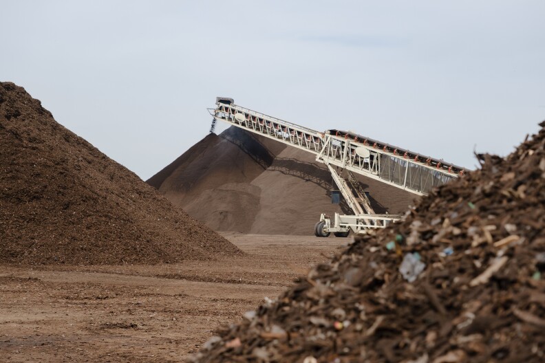 An image featuring mounds of compost, a pile of green waste in the foreground. A large crane dumps compost onto a pile in the background. 