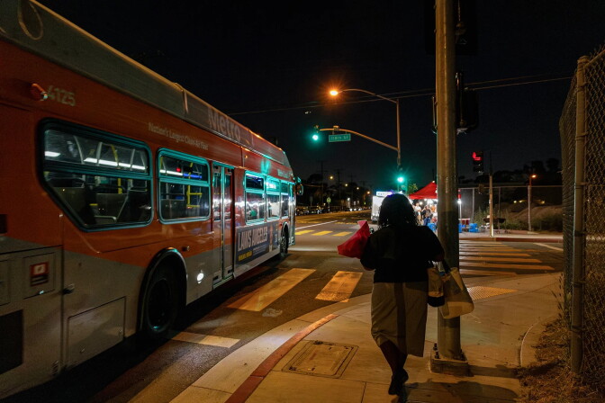 A woman is pictured from behind, at night, walking past an L.A. metro bus. She is carrying various bags and packages in her arms.