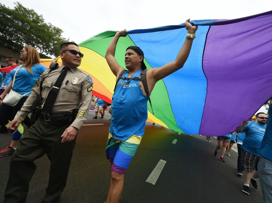 A person helps carry a large rainbow flag over their head while they march down the street next to a sheriff's deputy.