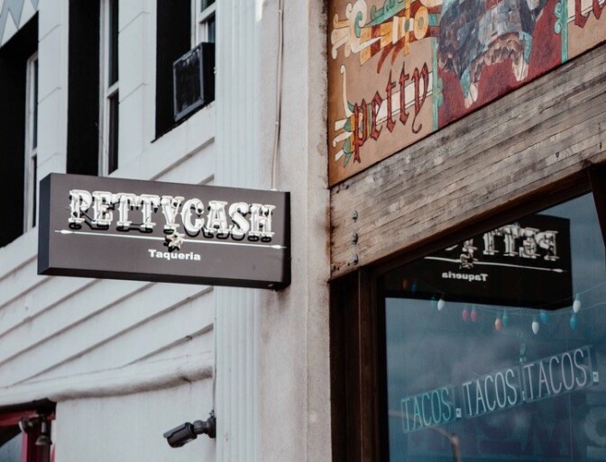 A sign hanging off the side of a rustic building reads "Petty Cash Taqueria," while another sign says "Tacos Tacos Tacos" through the window.