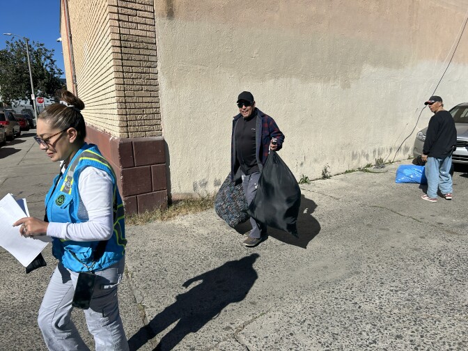 A man wearing black and holding two bags walks through an alleyway, following a woman wearing a blue vest. 