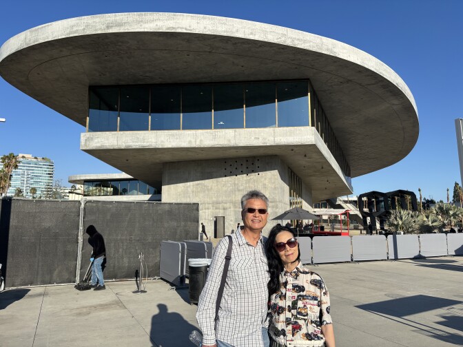 A man with gray hair, a plaid shirt and sunglasses next to a woman with black hair wearing sunglasses and a printed button down in front of temporary fences in front of the concrete and glass LACMA building. A clear blue sky in the background.