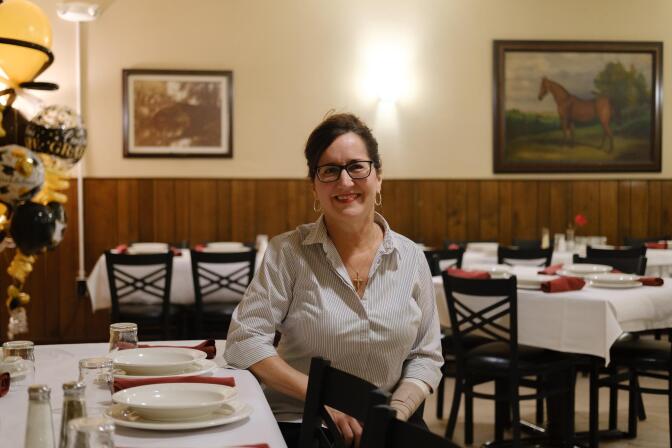 a woman in a white shirt sits next to a table covered in a white tablecloth with clean dishes waiting. she is in a semi-formal dining room