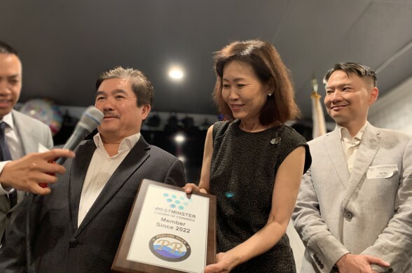 An Asian American woman in her 60s wearing a black shift dress presents a plaque from the Westminster Chamber of Commerce to an Asian American man in a suit.