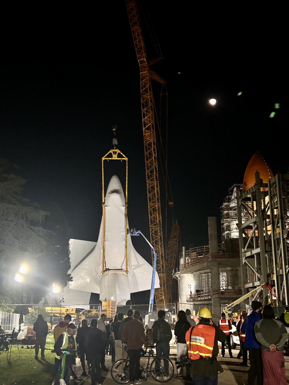 A group of scattered onlookers watch a huge, white space shuttle being lifted vertically by an even bigger yellow crane. A chain link fence separates the crowd from the crews.